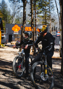 Two mountain bikers, an adult and a child, take a break at Howler Bike Park in Walnut Shade, MO. The park offers family-friendly trails for all ages and skill levels, promoting adventure, physical exercise, and quality bonding time. This scene captures the joy and togetherness that mountain biking brings to families.