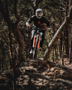 Mountain biker in full gear navigating a rocky trail through trees at Howler Bike Park during fall, equipped with a helmet, goggles, and layered clothing for cooler weather. The biker rides a bike with lower tire pressure for better traction on slick surfaces, as discussed in fall riding preparation tips.