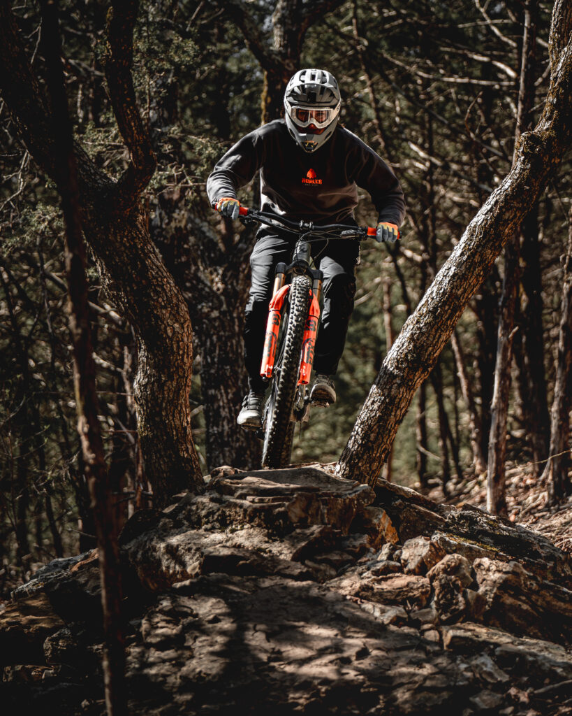 Mountain biker in full gear navigating a rocky trail through trees at Howler Bike Park during fall, equipped with a helmet, goggles, and layered clothing for cooler weather. The biker rides a bike with lower tire pressure for better traction on slick surfaces, as discussed in fall riding preparation tips.