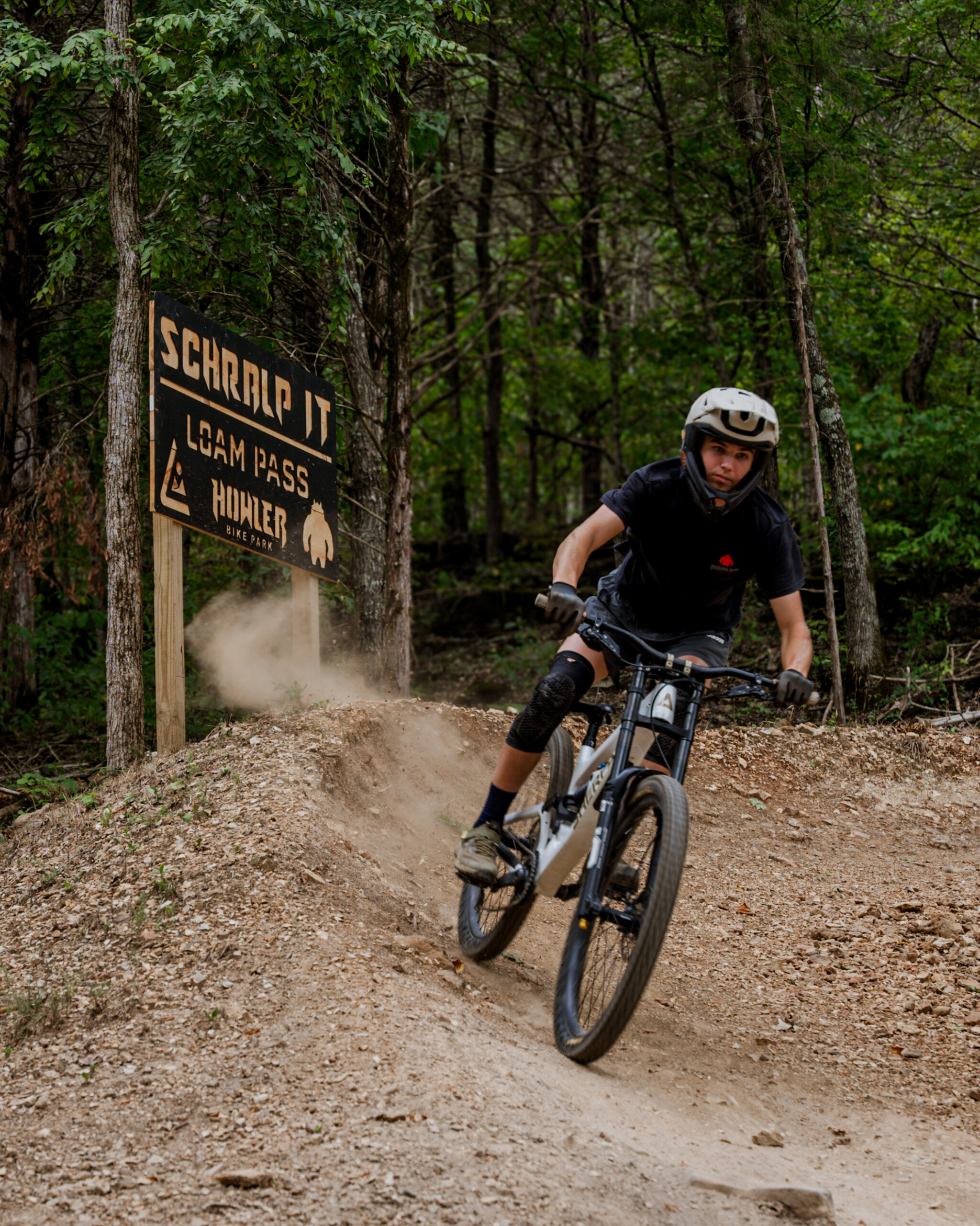 Mountain biker navigating a dirt trail at Howler Bike Park in the fall. The rider is leaning into a turn with a backdrop of dense trees, wearing safety gear including a helmet and knee pads. A trail sign reading 'Schralp It - Loam Pass' can be seen in the background, showcasing the perfect fall setting for an adventurous fitness routine.