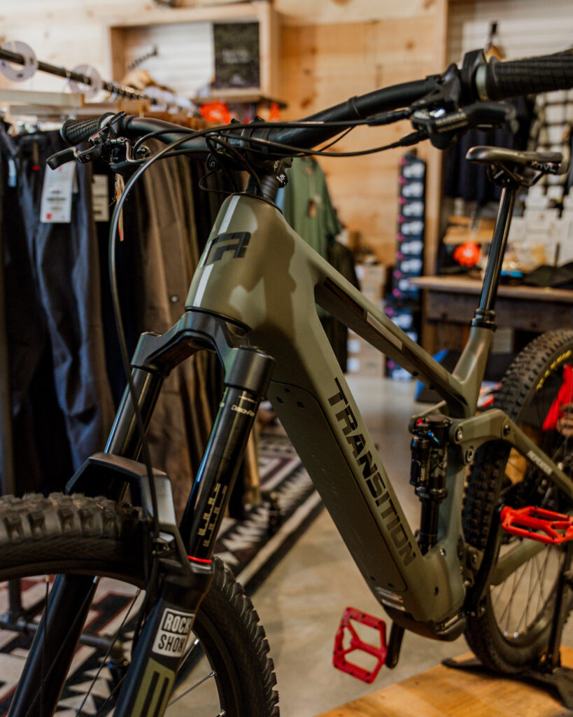 A Transition mountain bike on display in a shop, featuring a RockShox front suspension fork and red pedals. The bike is designed for trail and enduro riding, making it suitable for intermediate to advanced riders seeking a versatile, high-performance mountain bike. The shop in the background has various biking gear and clothing available for purchase.