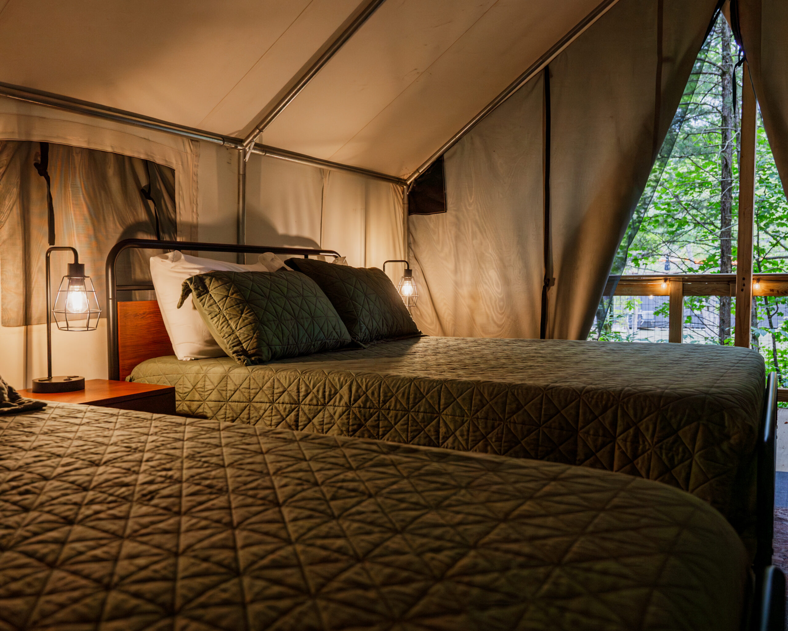 Interior view of a cozy glamping tent at Howler Bike Park in the fall, featuring two comfortable beds with quilted green bedding, soft ambient lighting, and a scenic view of the surrounding autumn forest through the open tent flaps. Perfect for an autumn outdoor adventure with a blend of comfort and nature immersion.