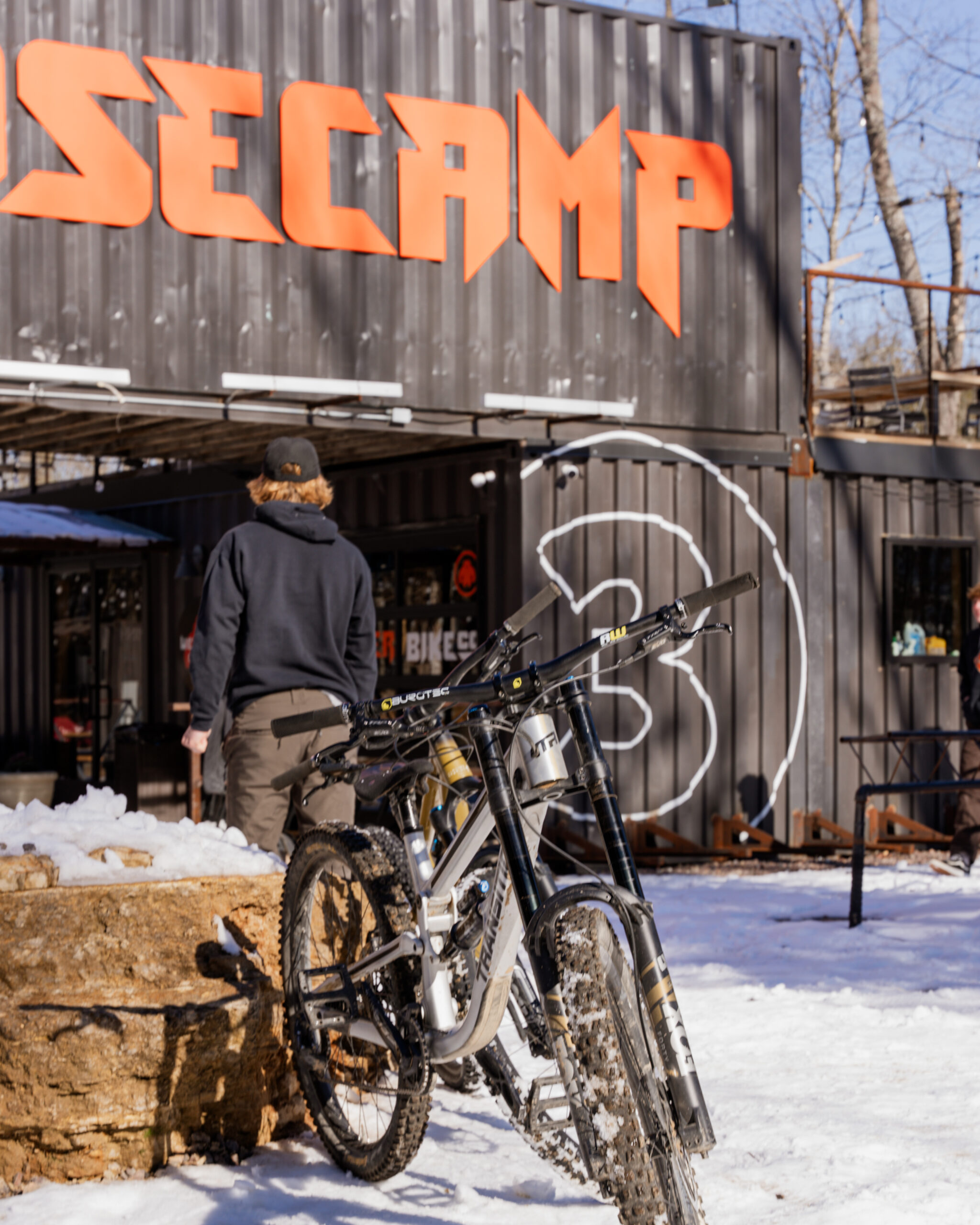 Mountain bikes with snow-covered tires parked outside Basecamp at Howler Bike Park during winter. A rider in cold-weather gear walks toward the entrance, showcasing off-season training for mountain bikers. The image highlights winter riding, fat biking, and maintaining endurance for the spring season.
