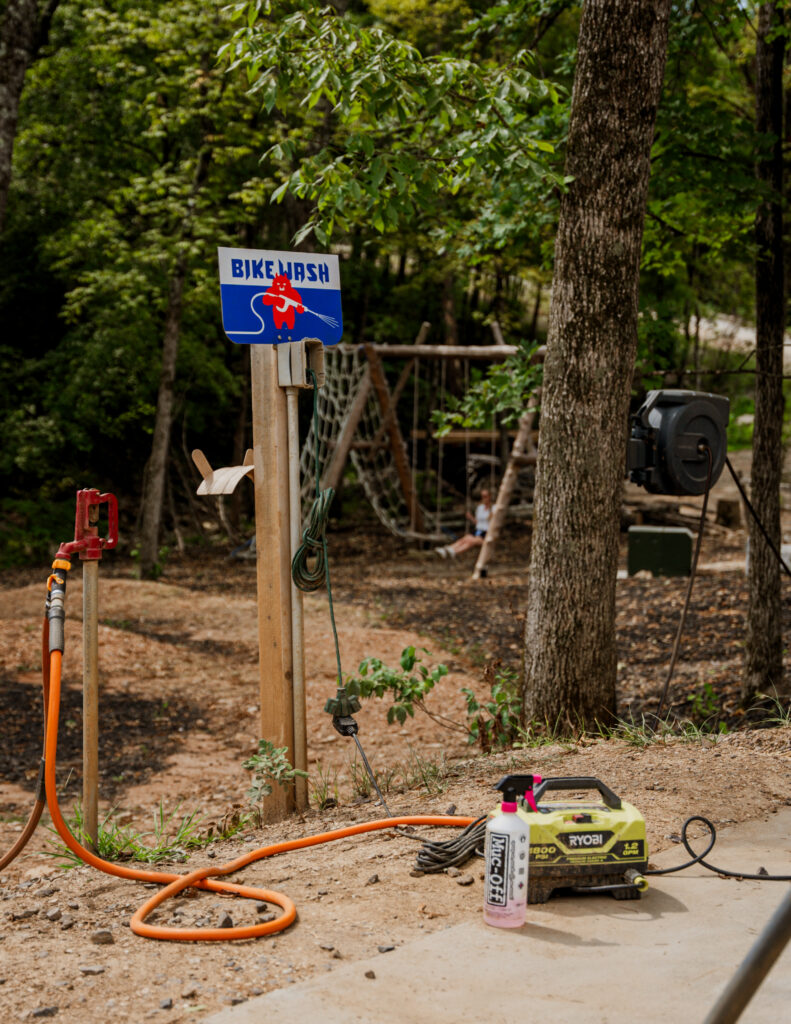 Bike wash station at Howler Bike Park featuring a Ryobi pressure washer, Muc-Off bike cleaner, and an orange hose, set against a wooded backdrop with a playground in the distance. A 'Bike Wash' sign with a red bear holding a hose is mounted on a wooden post. Perfect spot for cleaning off winter grime, lubing chains, and prepping your mountain bike for spring riding.