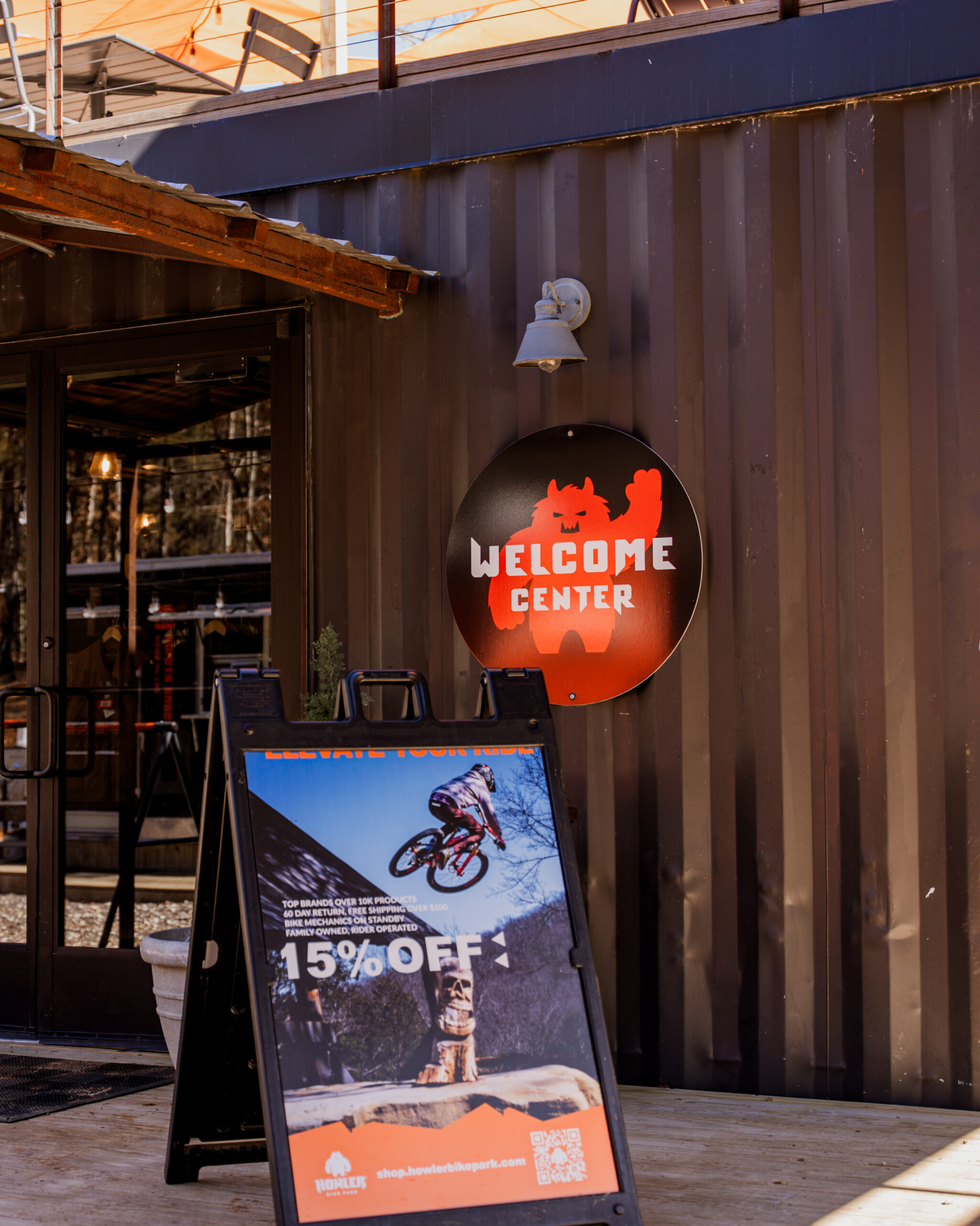 Howler Bike Park Welcome Center with a 15% off promotional sign featuring a mountain biker mid-air on a jump. The entrance is framed by a modern, industrial-style container building, with warm lighting inside. The sign encourages riders to gear up for spring break adventures, highlighting discounts on biking essentials. Perfect spring riding conditions, budget-friendly fun, and thrilling trails make Howler the ultimate alternative to a crowded beach getaway